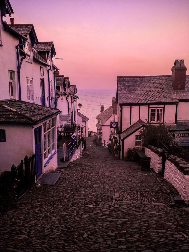 Dusk view down a steep old cobbled street lined with quaint cottages with the ocean in the distance