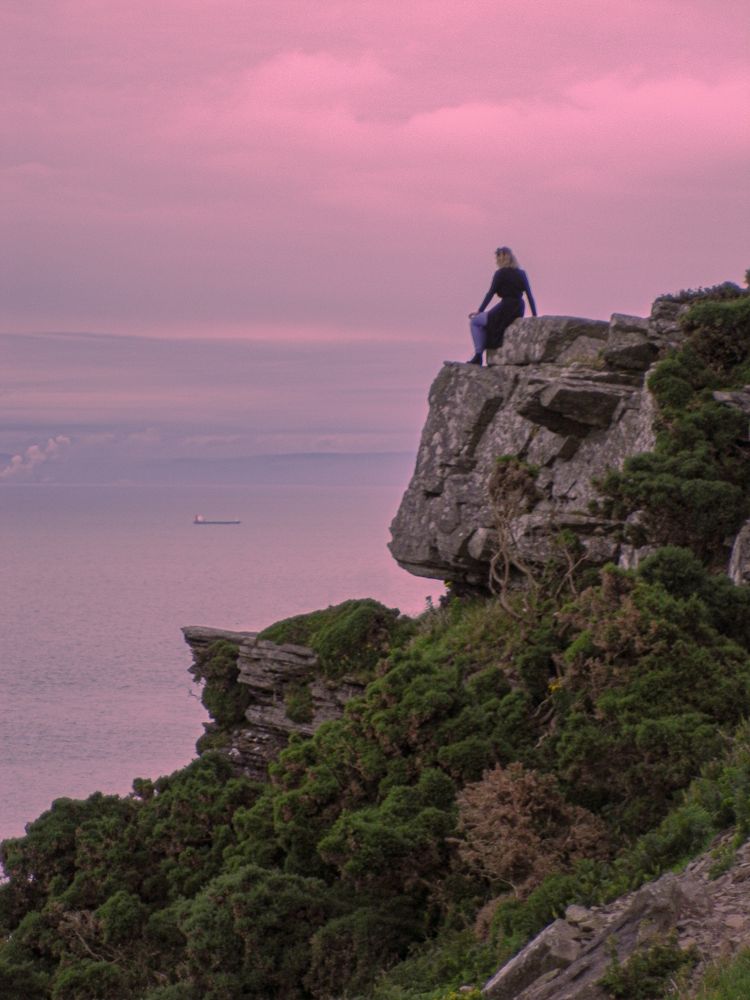 A woman sitting on clifftop rocks gazes out to sea towards a distant ship